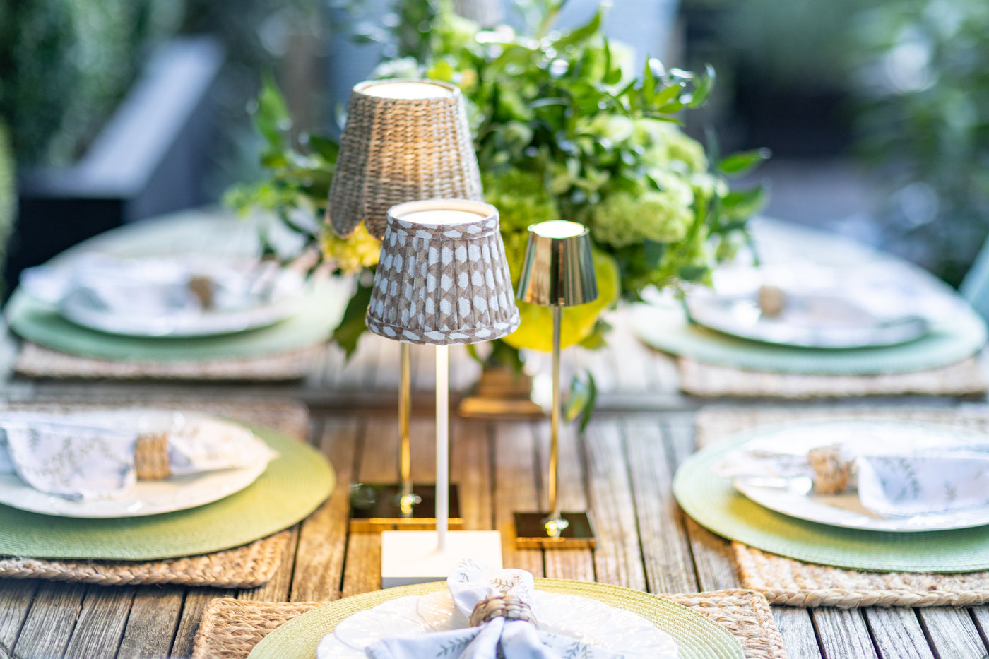 Outdoor dining table setting with Poldina cordless lamps, a seagrass shade, and Fermoie brown wicker shade, and a lush green floral centerpiece on a teak terrace table.