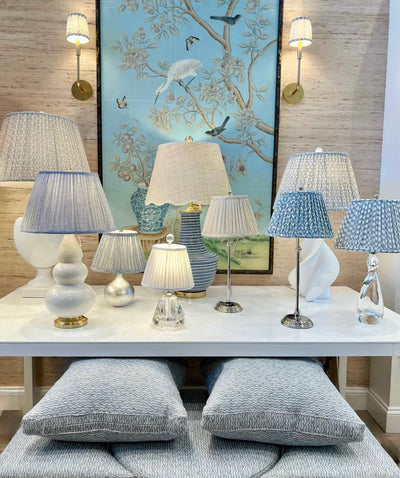 Display of luxury designer lampshades, including the Fermoie Blue Moire gathered empire lampshade on a white ceramic lamp base, alongside other blue and neutral-toned shades in a showroom setting.