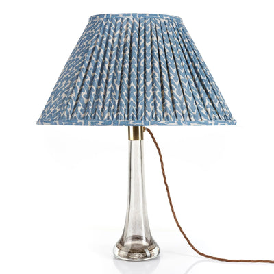 Front view of designer Fermoie Blue Rabanna gathered empire oval lampshade with pleated detailing displayed on a clear glass lamp base.