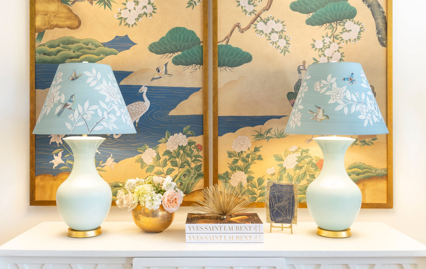 Decorative table setting with two lamps, a vase, and books against a wall with floral and landscape design.