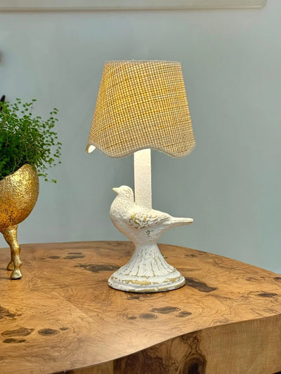 Decorative lamp with bird design on a wooden table next to a gold plant pot.