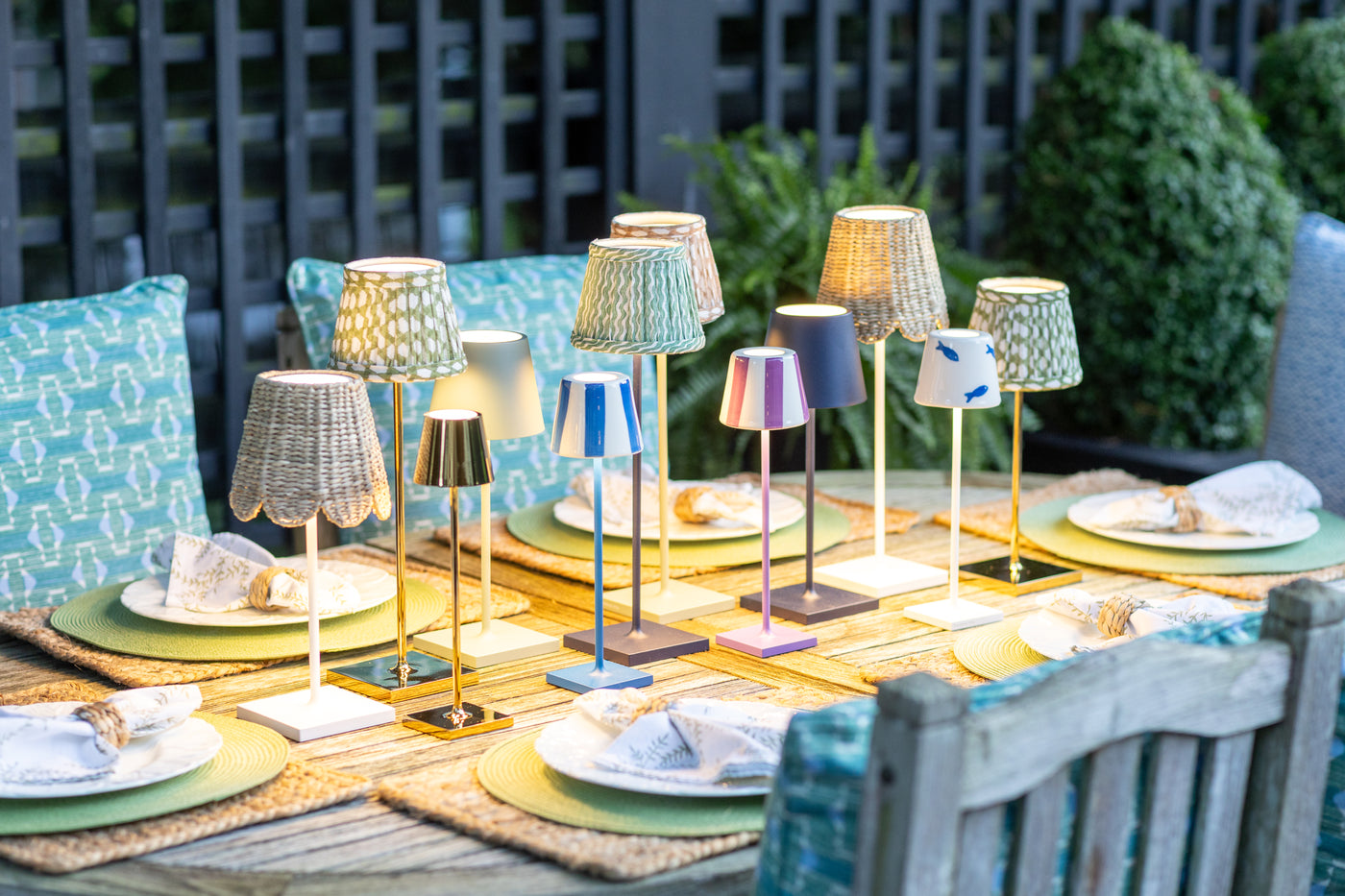 A styled outdoor dining table display featuring multiple small table lamps with patterned and solid shades in beige, blue, pink, and white, set against teal patterned cushions and greenery.