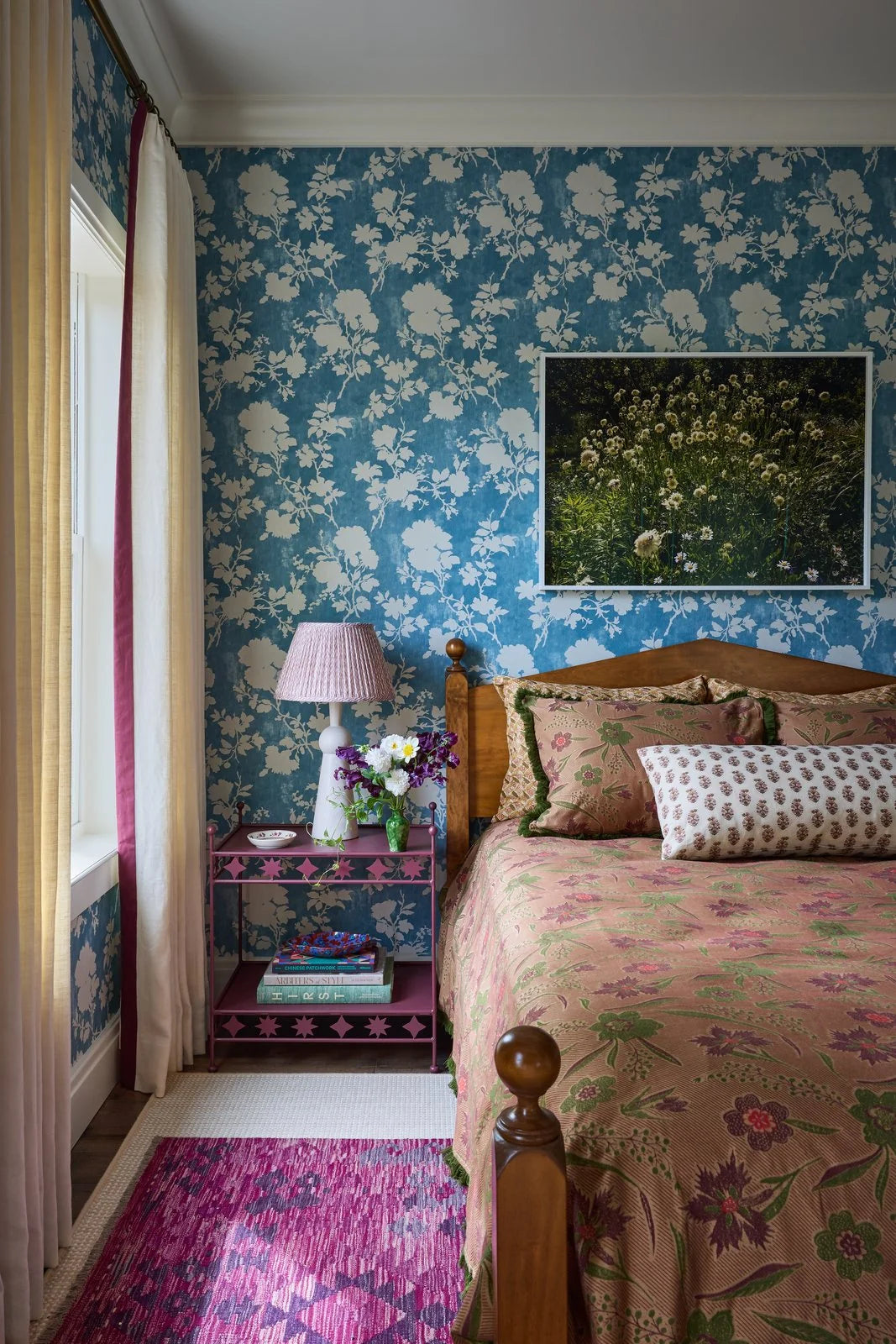 A pink pleated lampshade on a pale lamp beside a bed, with blue floral wallpaper, a pink nightstand, and a pink rug.