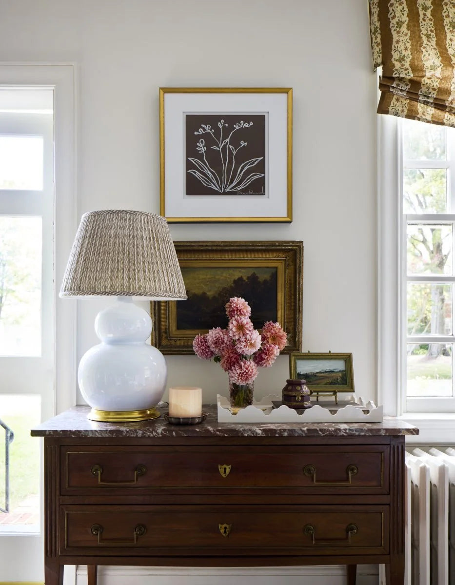 A pleated brown wave-stripe lampshade on a white gourd lamp beside a patterned Roman shade in Ashley Hanley’s room.