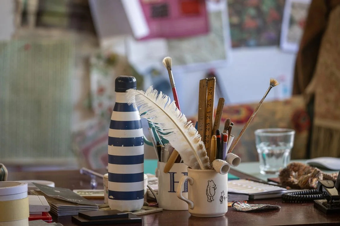 A mug of pencils, brushes, and a feather quill on the design table in front of a wall of pinned references