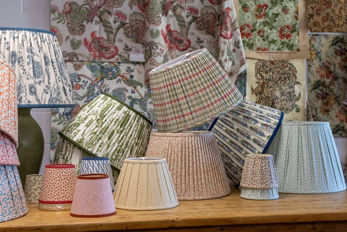 Pleated lampshades in plaids, stripes, and small prints stand on a wooden table in front of hanging floral fabrics