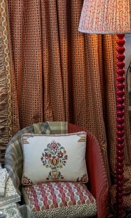 A pleated lampshade lit on a red bobbin floor lamp beside a wicker chair dressed in the collection’s prints